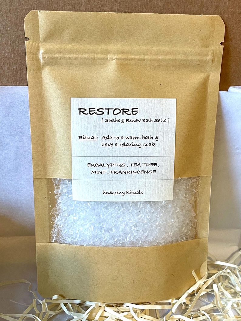 May include: A brown paper bag containing white bath salts, with a clear window. The label reads "RESTORE" and lists ingredients and instructions. The text "Unboxing Rituals" is also visible on the label.