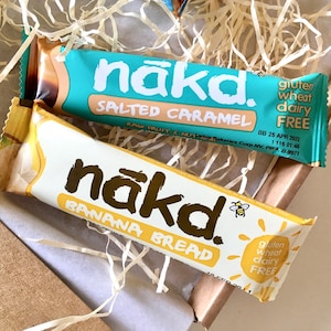 May include: Two rectangular snack bars labelled nākd. The top bar is turquoise with Salted Caramel text, and the bottom bar is white with Banana Bread text. Both wrappers feature the nākd logo, a small bee icon, and state gluten, wheat, and dairy free.