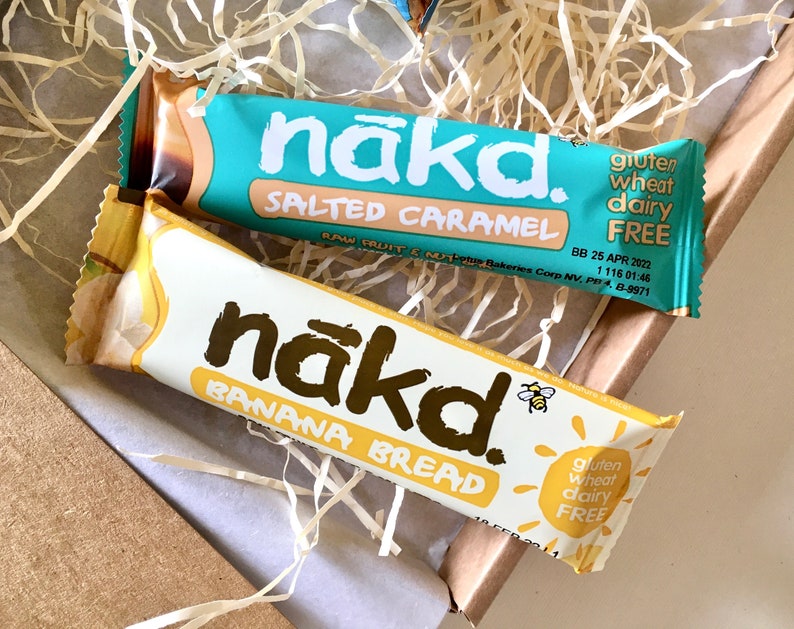 May include: Two rectangular snack bars labelled nākd. The top bar is turquoise with Salted Caramel text, and the bottom bar is white with Banana Bread text. Both wrappers feature the nākd logo, a small bee icon, and state gluten, wheat, and dairy free.