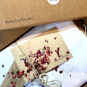 May include: A brown cardboard box with the words "Unboxing Rituals" printed on the top. The box is open and inside is a white tissue paper with a small brown card that says "Especially for you". The card is decorated with dried flower petals. There is a small wooden heart tied with twine on top of the tissue paper.