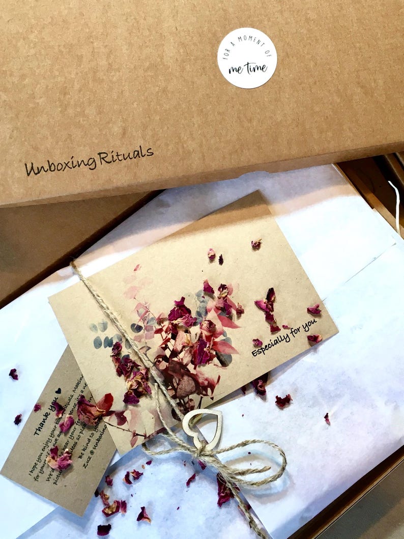 May include: A brown cardboard box with the words "Unboxing Rituals" printed on the top. The box is open and inside is a white tissue paper with a small brown card that says "Especially for you". The card is decorated with dried flower petals. There is a small wooden heart tied with twine on top of the tissue paper.