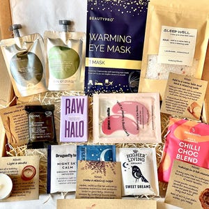 May include: A gift box filled with self-care items, including bath salts, tea bags, eye masks, and chocolate. The box is filled with shredded paper and has a variety of colours and textures. The text on the items includes "Sleep Well", "Warming Eye Mask", "Night Sky Calm", "Higher Living", "Sweet Dreams", and "Chilli Choc Blend".
