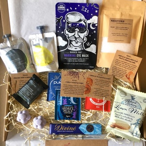 May include: A gift box filled with various self-care items, including bath salts, tea bags, eye masks, bath bombs, chocolate, and a lemon melt treat. The box is filled with shredded paper and has a brown cardboard exterior.