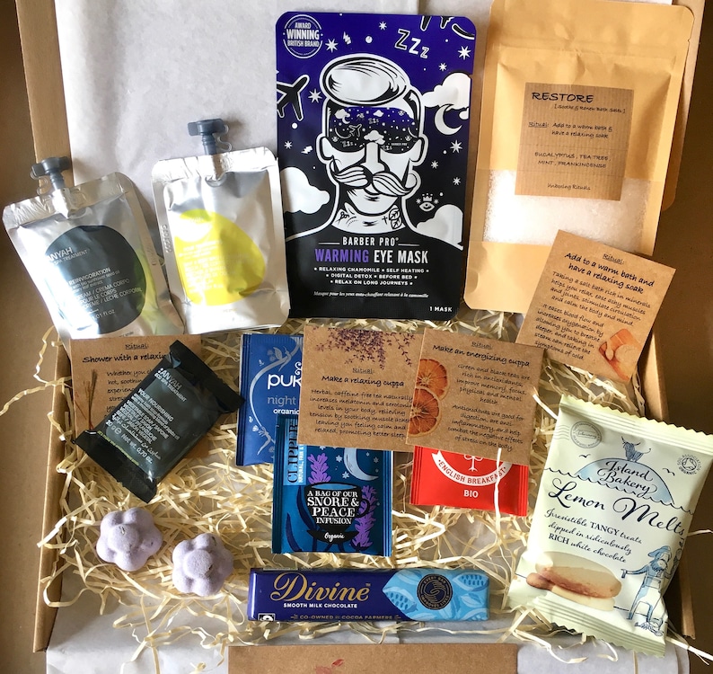 May include: A gift box filled with various self-care items, including bath salts, tea bags, eye masks, bath bombs, chocolate, and a lemon melt treat. The box is filled with shredded paper and has a brown cardboard exterior.