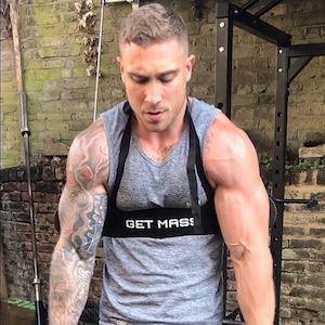 May include: A man wearing a gray tank top with the text "GET MASS" on a black strap, black shorts, and a black arm strap, is lifting two black dumbbells.