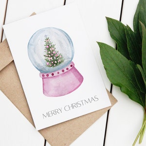 May include: A white card with a watercolour illustration of a pink snow globe with a green Christmas tree inside. The text "Merry Christmas" is printed on the card.