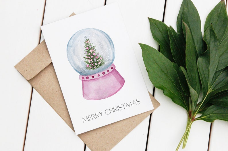 May include: A white card with a watercolour illustration of a pink snow globe with a green Christmas tree inside. The text "Merry Christmas" is printed on the card.