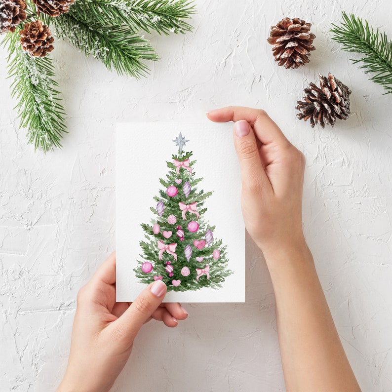 May include: A watercolor Christmas tree illustration on a white card. The tree is decorated with pink bows, ornaments, and a silver star on top. The card is held against a white textured background with pine needles and pine cones.