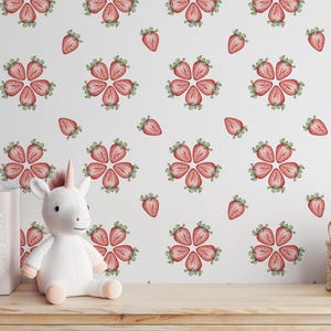 May include: A white stuffed unicorn toy sits in front of a white wall with a pink and green strawberry pattern. The wall is covered in a repeating pattern of strawberries.
