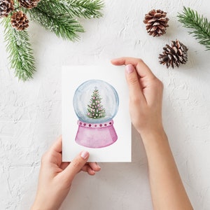 May include: A white card featuring a pink and white snow globe with a green Christmas tree inside. The snow globe is set against a white background.