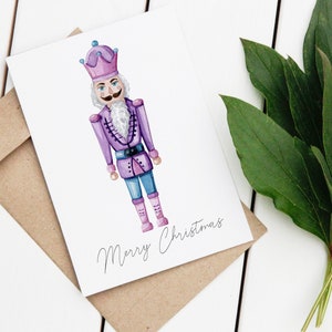 May include: A watercolor Christmas card featuring a purple nutcracker soldier with a white beard and mustache. The card is on a white surface with a brown envelope and green leaves. The words "Merry Christmas" are written on the card.