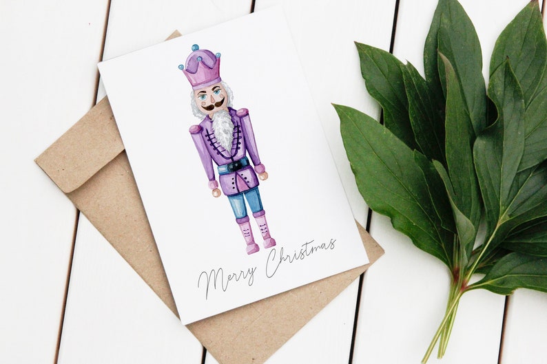 May include: A watercolor Christmas card featuring a purple nutcracker soldier with a white beard and mustache. The card is on a white surface with a brown envelope and green leaves. The words "Merry Christmas" are written on the card.
