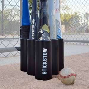 May include: A black cylindrical baseball bat rack with the white text STICKSTOW printed on the front. The rack holds five baseball bats of various colors and brands, including Easton and Louisville, next to a worn white baseball with red stitching.