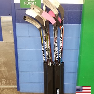 May include: A black hockey stick holder with four slots, holding multiple hockey sticks with the brand names "Bauer" and "CCM" printed on them. The holder is labeled "Stickstow" and is mounted on a wall with a blue and white brick pattern. A sign above the holder reads "Team Room 1".