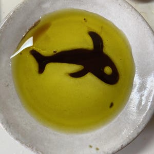May include: A round, off-white ceramic bowl filled with bright yellow olive oil. Floating in the center of the oil is a dark brown balsamic vinegar reduction shaped like an orca whale, featuring a distinct dorsal fin and a small circular eye.