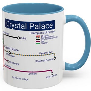 May include: A white ceramic mug with a blue interior and handle. The mug features a graphic of a subway map with the words "Crystal Palace" and "Champions of Europe." The map includes team statistics and various city names.