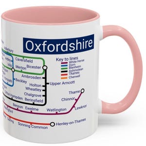 Peut inclure: Une tasse en céramique blanche avec un bord et une poignée roses. La tasse présente une carte de l'Oxfordshire, en Angleterre, avec une légende des différentes lignes de train. La carte comprend les noms des villes et des villages de la région.
