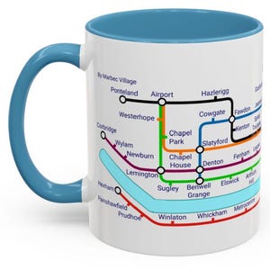 Puede incluir: Taza de cer&aacute;mica blanca con borde y asa azules. La taza presenta un mapa estilizado del sistema de metro de una ciudad con nombres de estaciones y l&iacute;neas de colores.