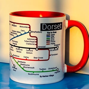 May include: White ceramic mug with a red rim and handle. The mug features a map of Dorset, England, with various towns and cities labelled. The map is styled like a London Underground map, with different coloured lines representing different routes. The text "Dorset" is printed in large, bold letters at the top of the mug. The text "Key to lines" is printed below the map, with a legend explaining the different coloured lines.