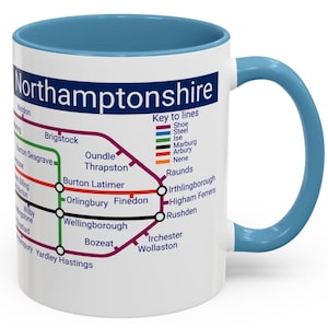 May include: White ceramic mug with a blue rim and handle. The mug features a map of Northamptonshire, England, with various train lines and station names. The map is in a vintage style with colourful lines and black circles for stations.