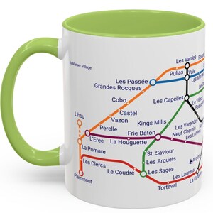 Puede incluir: Una taza de cer&aacute;mica blanca con un borde y un asa verdes. La taza presenta un mapa estilizado del metro de una ciudad con varias l&iacute;neas de colores y nombres de estaciones. El mapa incluye los nombres de las estaciones como "Les Pass&eacute;e", "Grandes Rocques", "Cobo", "Lihou", "Perelle", "L'Eree", "La Pomare", "Les Clercs", "Pleinmont", "Le Coudr&eacute;", "Kings Mills", "Frie Baton", "La Houguette", "Les Capelles", "Pulias", "Vale", "Les Vardes", "Le Village", "Beauc", "Les Varendes", "Neuf Chemin", "Les Lohiers", "St. Saviour", "Les Arquets", "Les Sages", "Les Laurens", "Torteval" y "La Corb".