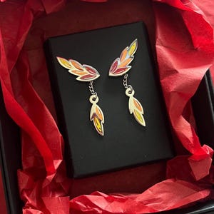 May include: A pair of gold-toned metal earrings featuring a wing-shaped stud connected by a small chain to a dangling leaf-shaped charm. The earrings are decorated with enamel in shades of yellow, orange, and red, displayed inside a black box.