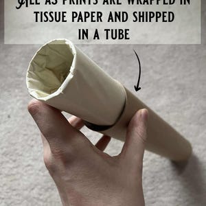 May include: A brown cardboard tube with a white paper lining. The tube is being held by a hand. The text "ALL A3 PRINTS ARE WRAPPED IN TISSUE PAPER AND SHIPPED IN A TUBE" is visible in the background.