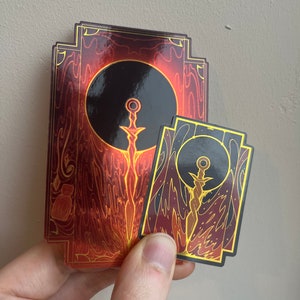 May include: Two gold and red stickers with a black background and a gold sword design. The larger sticker has a circular design with the sword in the center. The smaller sticker has a more rectangular design with the sword in the center.