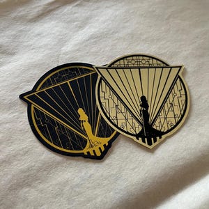 Puede incluir: Dos pegatinas circulares doradas y negras con un diseño geométrico y una silueta de una persona de pie en el centro. Las pegatinas están superpuestas.