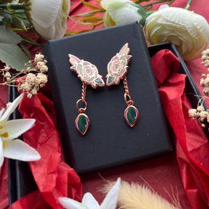 Könnte beinhalten: Ohrringe aus Roségold mit floralem Design und grünen tropfenförmigen Anhängern. Die Ohrringe werden in einer schwarzen Schachtel auf rotem Untergrund präsentiert, umgeben von weißen Blumen und rotem Seidenpapier. Die Ohrringe haben eine zarte, feminine Ästhetik.