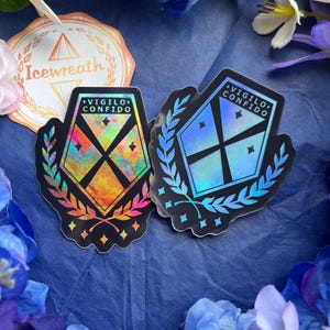 May include: Two shield-shaped stickers featuring a black cross design, laurel leaf accents, and the text VIGILO CONFIDO. One sticker displays a multi-colored holographic finish, while the other features a cool-toned blue holographic effect.
