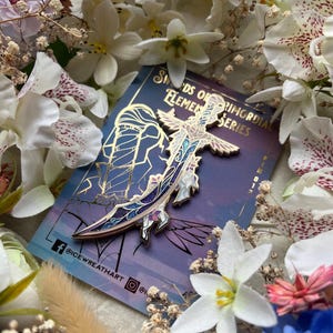 Op de afbeelding: Een goudkleurige emaille speld in de vorm van een gebogen zwaard met ingewikkelde blauwe, paarse en witte bloemmotieven. De speld is bevestigd op een rechthoekige kaart met de tekst Swords of Primordial Elemental Series en Icewreathart.