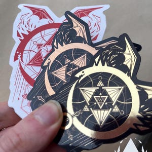 Similar to the previous photo, a hand holds the three variants of the sticker, but at a slight angle, to show them reflected by light. Icewreath's shop logo is placed in the corner.