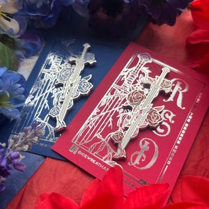 May include: Two silver-toned metal enamel pins shaped like swords entwined with roses, displayed on rectangular backing cards. One card is dark blue and the other is red, both featuring silver foil line art and the text @ICEWREATHAR.
