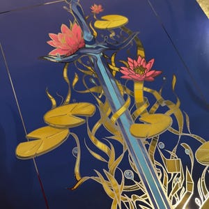 May include: A blue sword with a silver hilt is surrounded by gold and pink water lilies on a dark blue background.