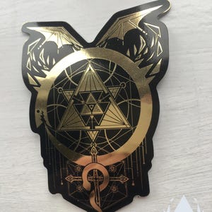 The Mirror Gold sticker sits at a slight angle on a white painted background, a light shining on the golden parts of the sticker, showing their reflective golden quality. Icewreath's shop logo is placed in the corner.