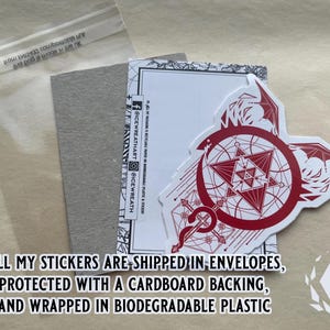 The Red sticker is displayed alongside the cardboard and biodegradable packaging used to ship the product. Text reads "All my stickers are shipped in envelopes, protected with a cardboard backing, and wrapped in biodegradable plastic."