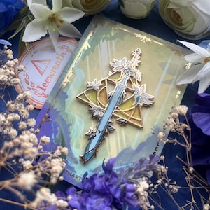 Peut inclure: Une broche en émail métallique représentant une épée bleu clair centrée dans un cadre triangulaire doré. L'épée est ornée de détails floraux blancs et bleus. La broche est fixée sur une carte de support rectangulaire avec le texte Icewreath.