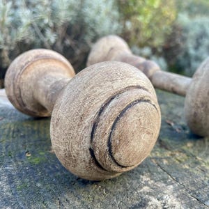 May include: Three antique wooden door knobs with a weathered, light brown finish. The knobs have a rounded shape with carved circular details. The background shows greenery and a wooden surface.
