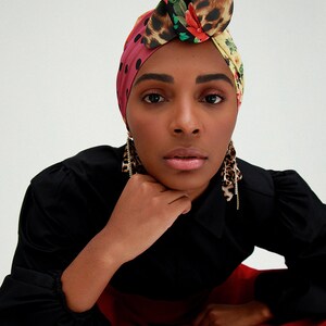 May include: A woman wearing a black shirt and red pants with a colorful patterned headscarf and gold earrings.
