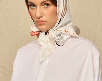 May include: A white silk scarf with a black and orange abstract print. The scarf is tied around the head of a person with brown hair.