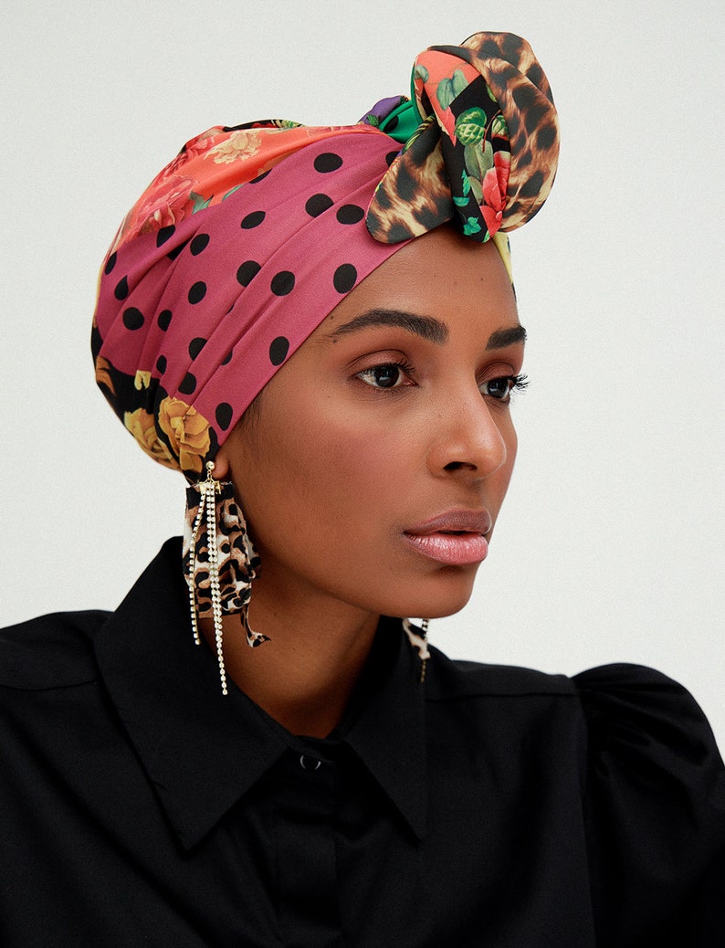 May include: A woman wearing a black shirt with a colorful patterned headscarf tied in a bow. The headscarf features polka dots, floral designs, and animal print. She is wearing large, dangling earrings with a leopard print design.