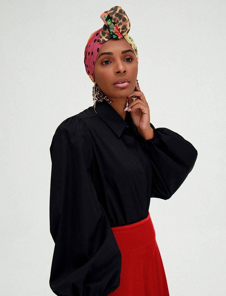 May include: A woman wearing a black blouse with long sleeves and a red skirt. She is wearing a colorful patterned headscarf with a leopard print design.