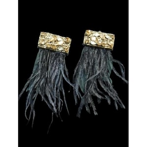 May include: A pair of dangling earrings with a rectangular gold-colored top adorned with clear and gold-toned stones. Below, long, dark gray feathers cascade downward, creating a dramatic and elegant look. The earrings are set against a black background.