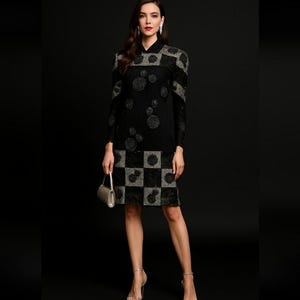 Pode incluir: Um vestido preto com mangas compridas e bainha até ao joelho. O vestido apresenta um padrão de quadrados prateados e pretos na bainha e enfeites florais prateados no corpete. A modelo usa saltos prateados e uma pequena bolsa prateada.