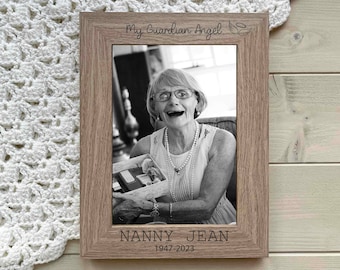 May include: A brown wooden picture frame with a black and white photo of a woman smiling. The frame has the text "My Guardian Angel" with a wing design and "NANNY JEAN 1947-2023" engraved on the bottom.