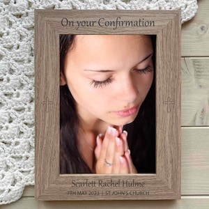 May include: A wooden photo frame with a light brown finish. The frame has a white mat and a photo of a person praying. The frame is engraved with the text "On your Confirmation", "Scarlett Rachel Hulme", "7TH MAY 2023", and "ST JOHN'S CHURCH".