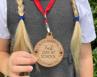 May include: A wooden medal with the text "Felicity Broomhall First Day At School 6th September 2022" and stars around the edge. The medal is hanging from a red ribbon around a person's neck.