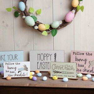 May include: Four wooden signs with Easter-themed messages. The signs are decorated in pastel colours and feature bunny silhouettes. The signs are arranged on a wooden surface with a wreath of pastel-coloured eggs and greenery in the background.