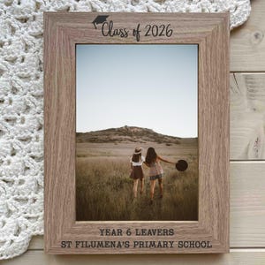 May include: A light brown wooden photo frame with the text "Class of 2026" and "Year 6 Leavers St Filumena's Primary School". The frame contains a photo of two people in a field.
